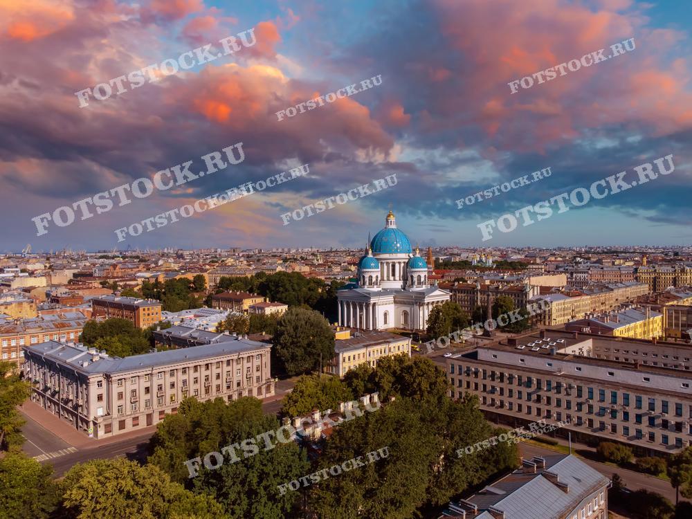На фотографии изображен Троицкий собор, окруженный городскими постройками, на фоне красочного закатного неба. Вечерние облака окрашены в розовые и оранжевые оттенки, создавая впечатляющий контраст с голубыми куполами собора. Снимок сделан с высоты, что позволяет оценить красоту и величие Санкт-Петербурга в белые ночи.